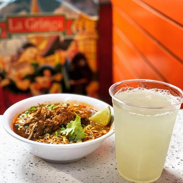 A bowl of birria ramen with a lime wedge and cilantro, next to a glass of lemonade.