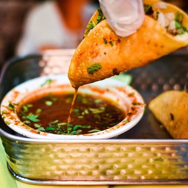 A hand dips a Tacos la Gringa birria taco into a bowl of consommé.