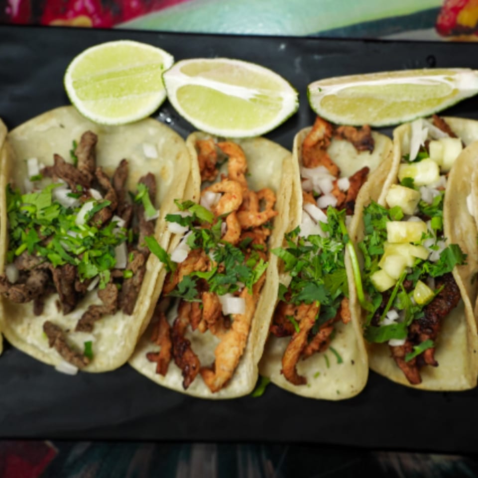 A black plate of Tacos la Gringa tacos with various fillings, cilantro, onions, and lime wedges.
