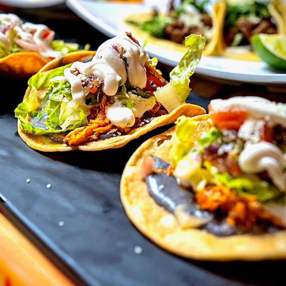 Close-up of three Tacos la Gringa tostadas with meat, lettuce, and crema on a dark surface.